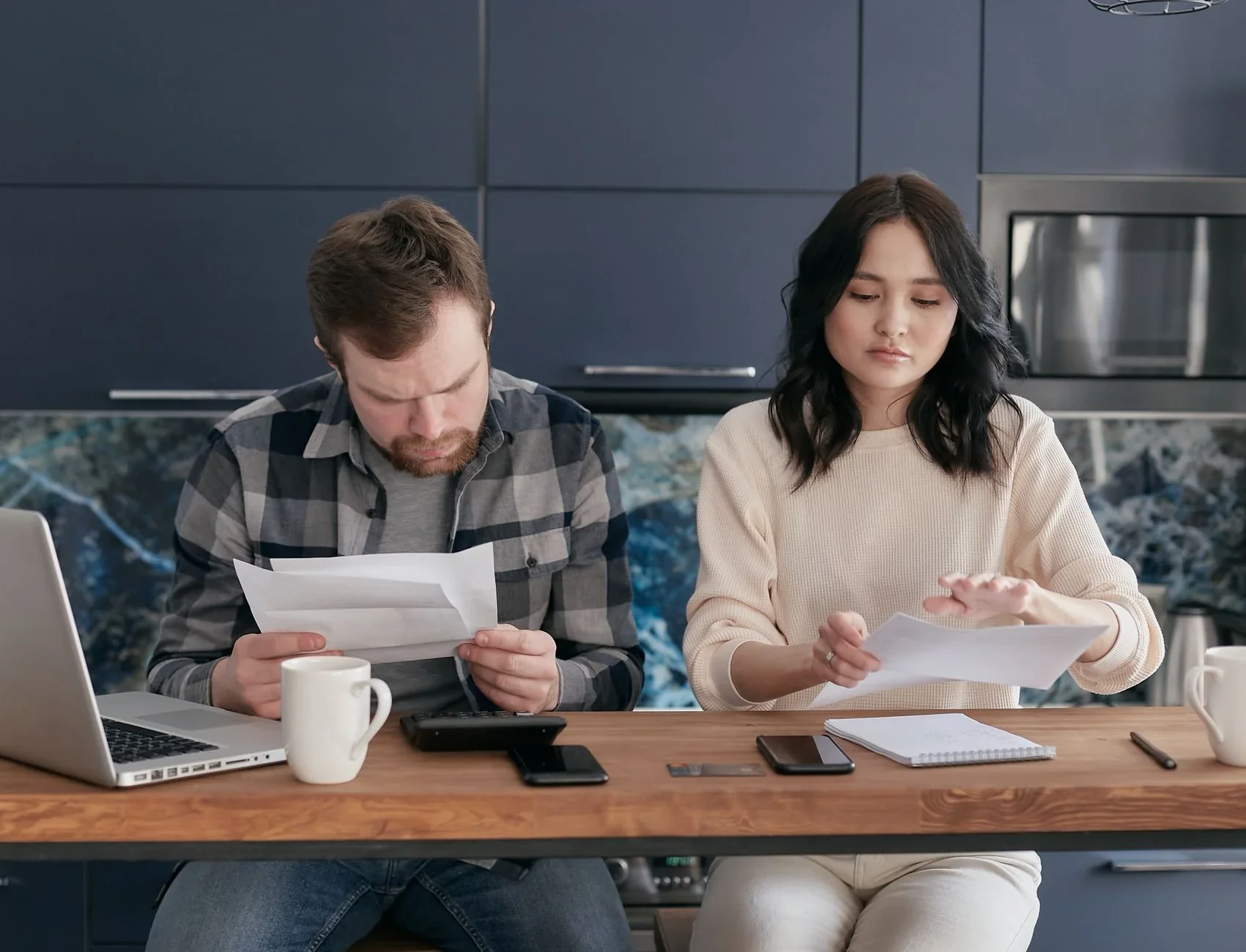 A couple at a kitchen table reviewing paper bills and notes beside a laptop, mug, calculator and notebook.