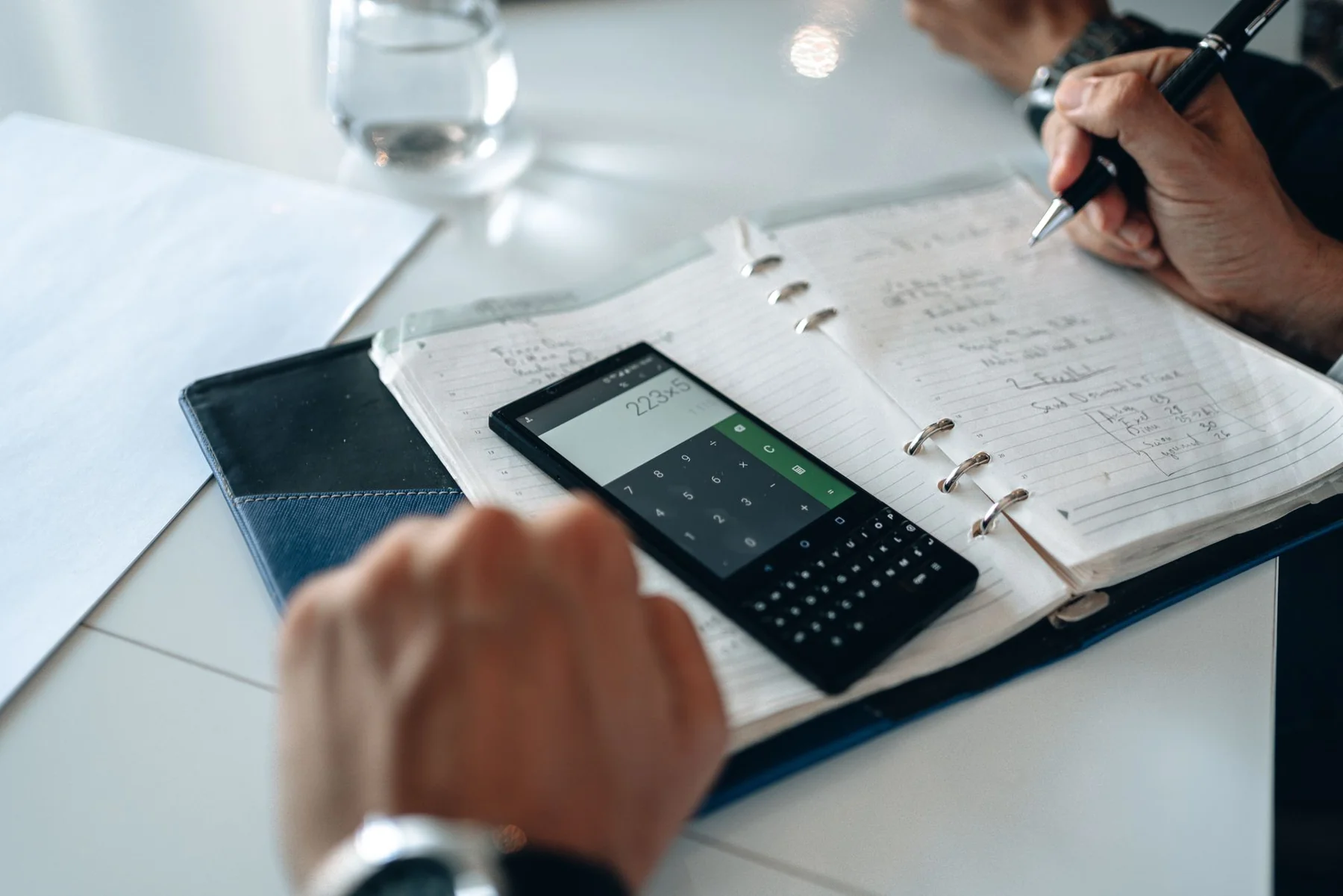 A person writing in a notebook beside a phone calculator.